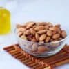Close-up of almonds in a glass bowl next to a bottle of oil on a bamboo mat.