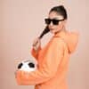 a woman in an orange hoodie holding a soccer ball