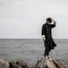Fashionable man in black coat and hat stands on rocky beach against ocean backdrop.