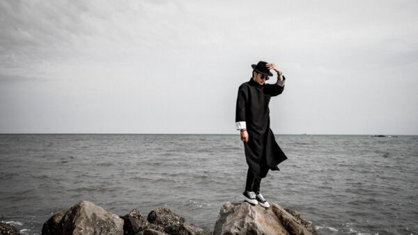 Fashionable man in black coat and hat stands on rocky beach against ocean backdrop.