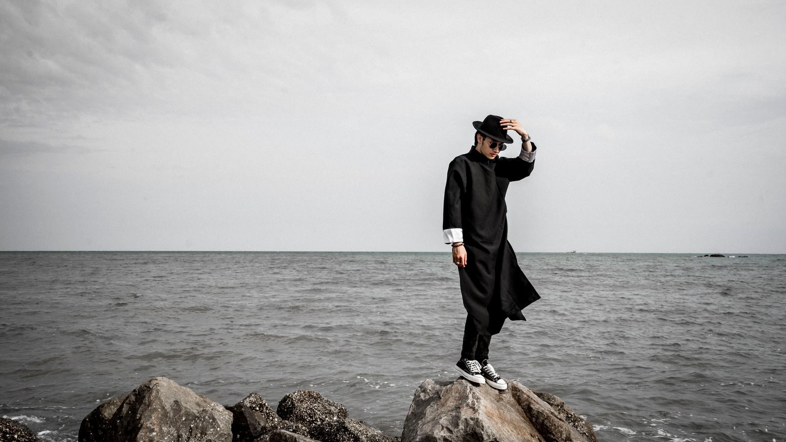 Fashionable man in black coat and hat stands on rocky beach against ocean backdrop.